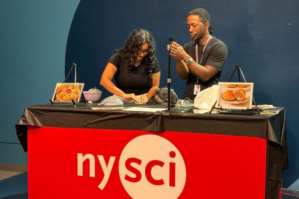Two educators, standing behind a table with the NySci logo, demonstrate how to fold dumplings.