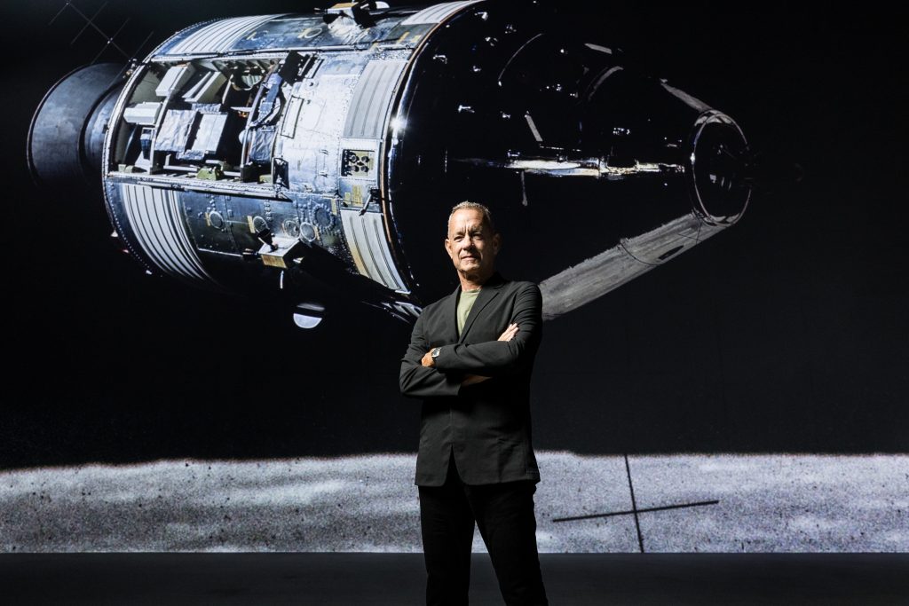 Photo of Tom Hanks in front of an historical image of an Apollo spacecraft