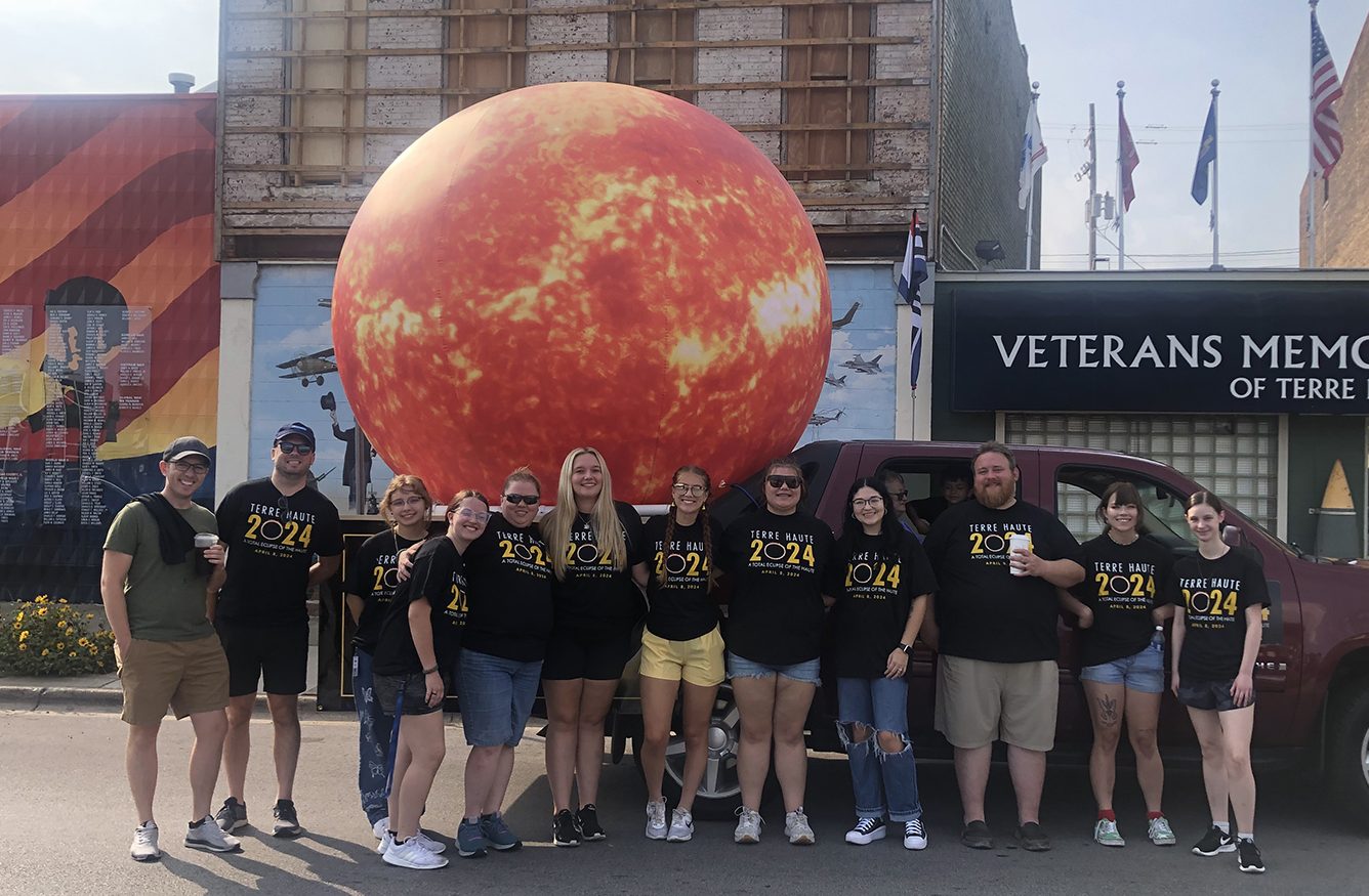Twelve people, all wearing black shirts that say "Total Eclipse 2024" stand in front of a parade float of a large inflatable sun.