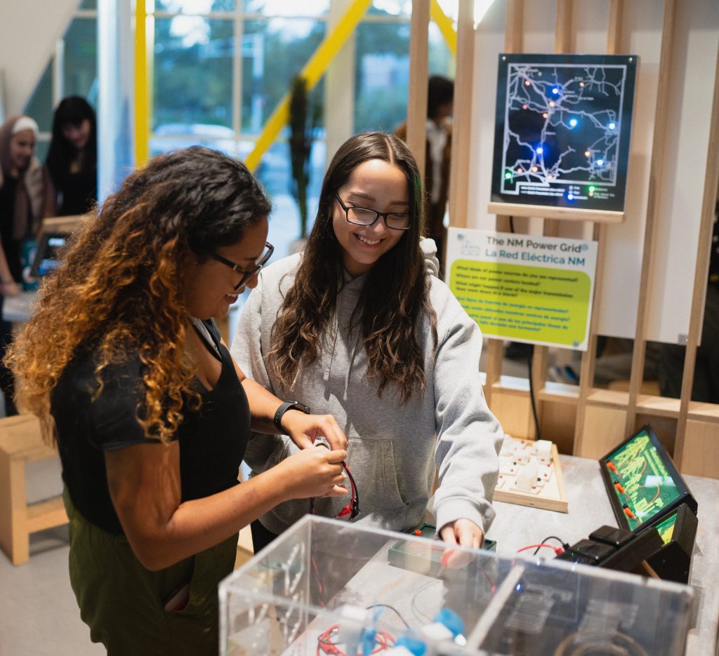 Two teen girls look together through a bin of electronics equipment.