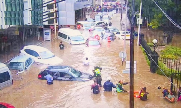 Muddy floodwaters fill a busy city street. About 20 cars are partially submerged and several people wade through the chest-deep water.