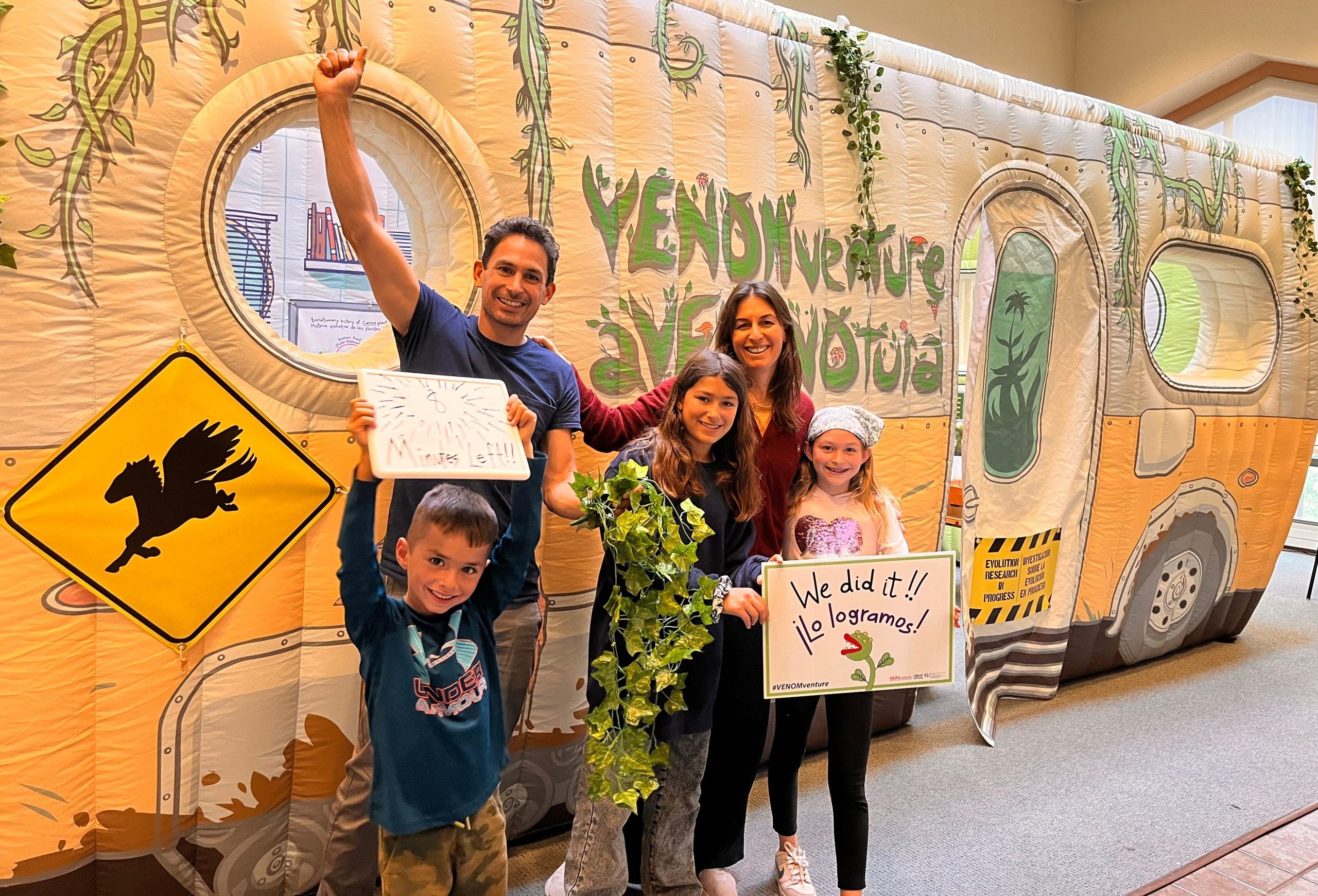 Two adults and three children strike a celebratory pose in front of the VENOMventure, inflatable escape room. They hold signs saying "We did it!" and "Lo logramos!"