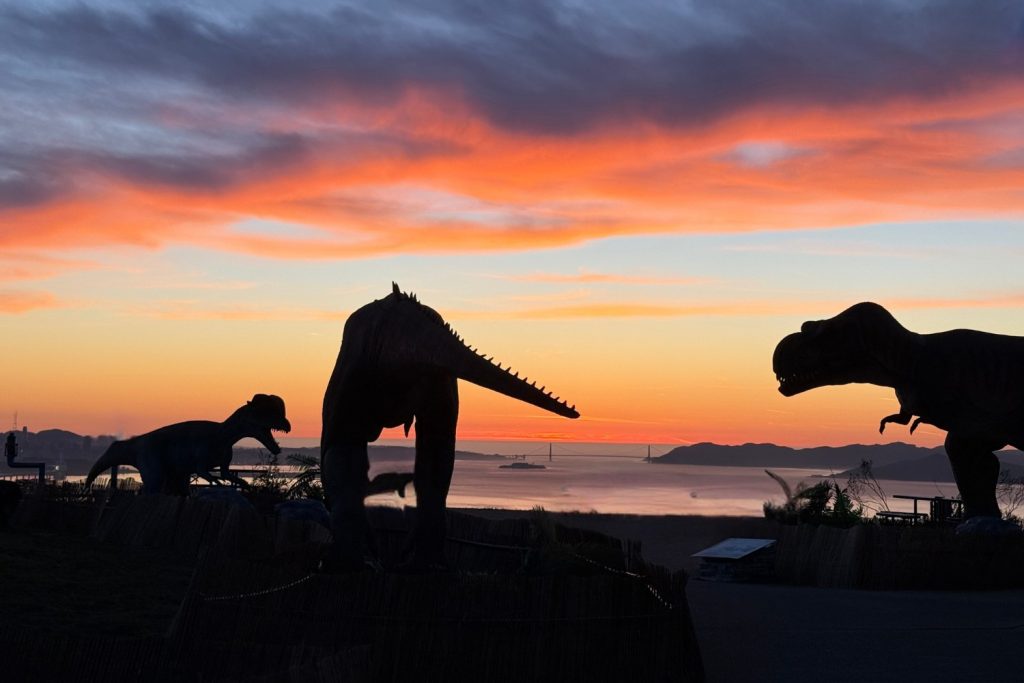 Three large dinosaur sculptures overlook the San Francisco Bay at sunset.
