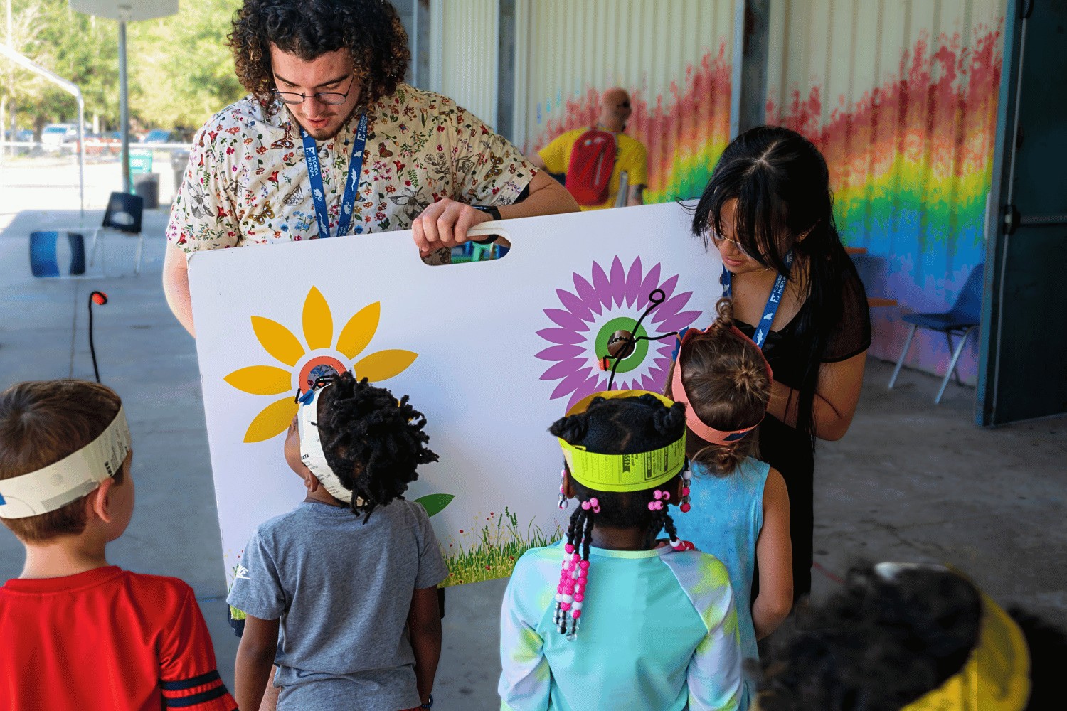 Two educators show a group of children an illustrated panel depicting two flowers. The children wear homemade pollinator hats.