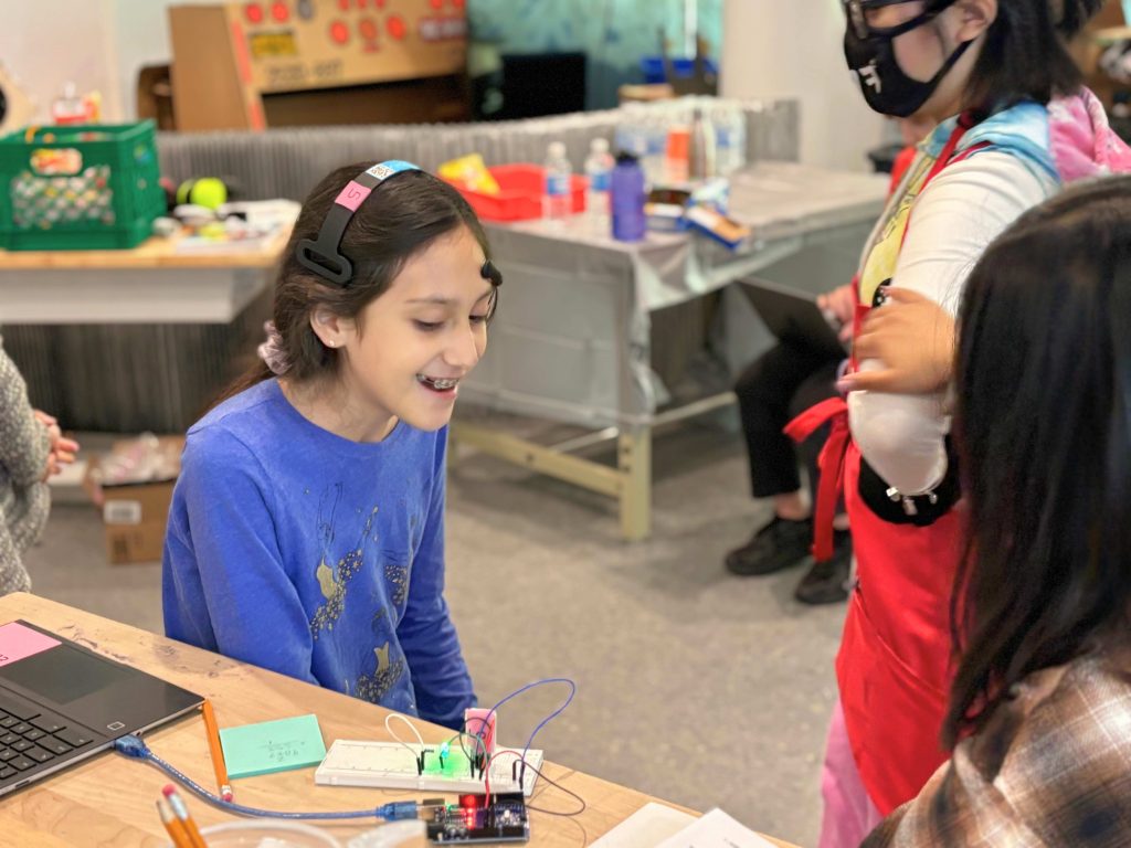 A smiling tween girl sits at a desk in a makerspace. In front of her is a laptop and circuit board. She is wearing a headset with electronic attachments.
