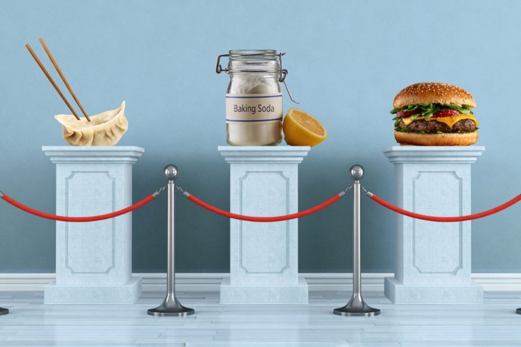 An image of a museum display with three pedestals, holding a dumpling, a jar of baking soda next to a lemon, and a cheeseburger.